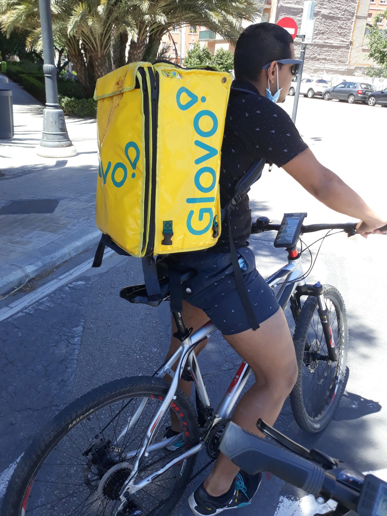 Repartidor de Glovo en bicicleta por una calle de Valencia con la mochila amarilla de la plataforma a la espalda.