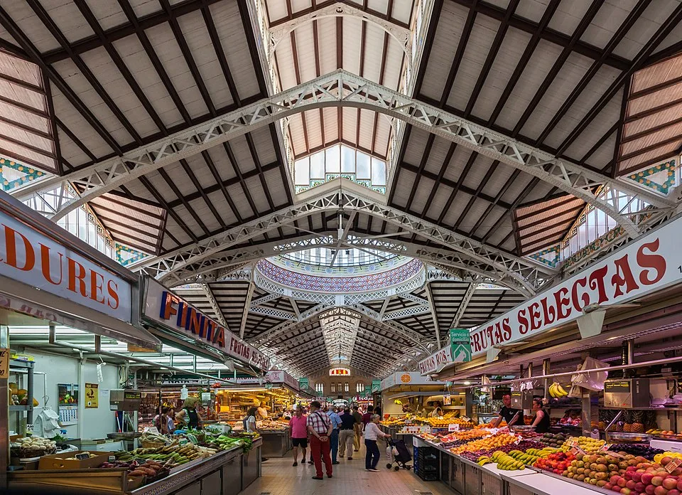 Vista interior del Mercado Central de Valencia, con puestos de frutas y verduras y compradores recorriendo el pasillo central.