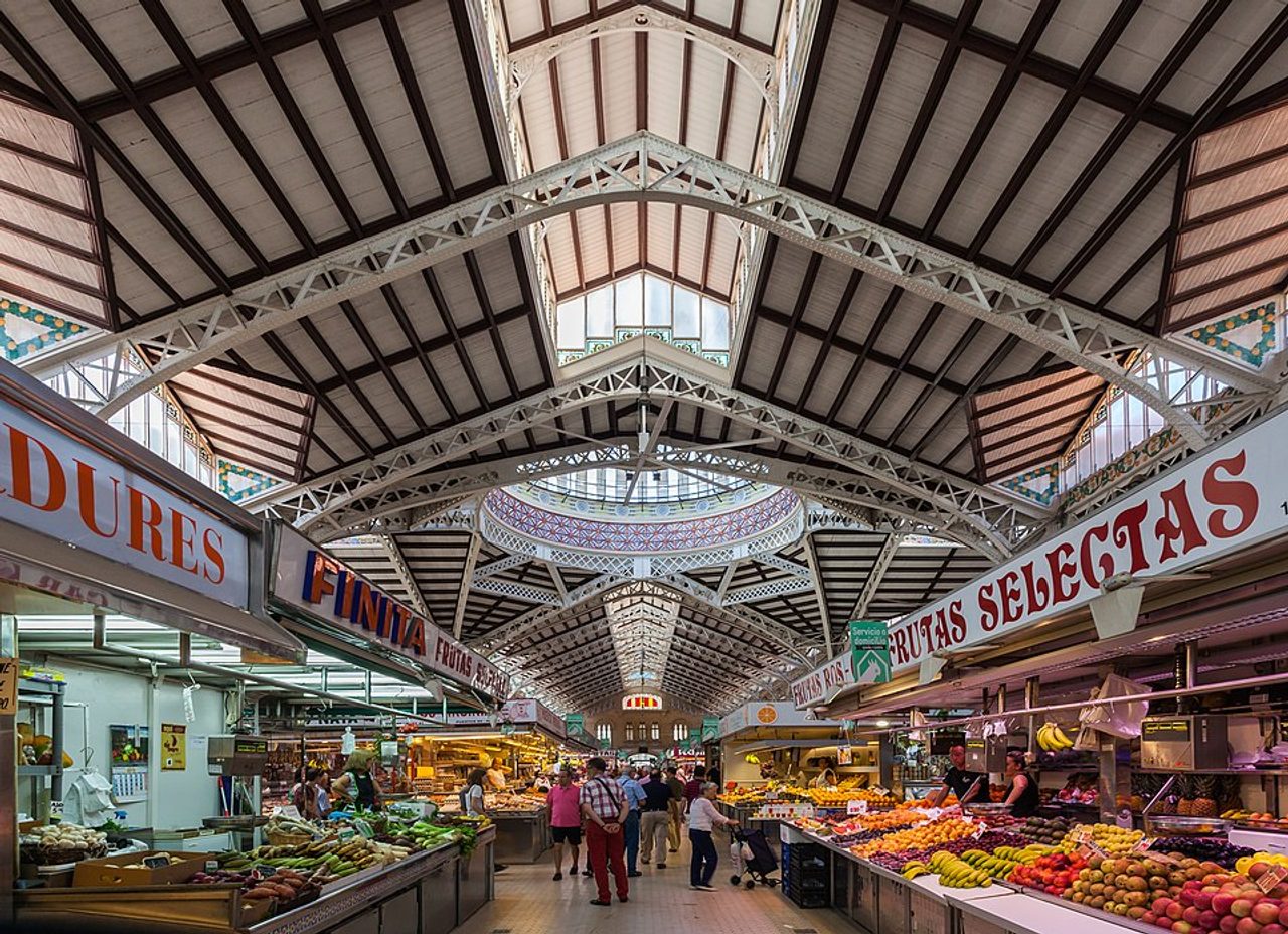 Interior del Mercado Central de Valencia, con puestos de frutas y verduras y compradores recorriendo el pasillo central.