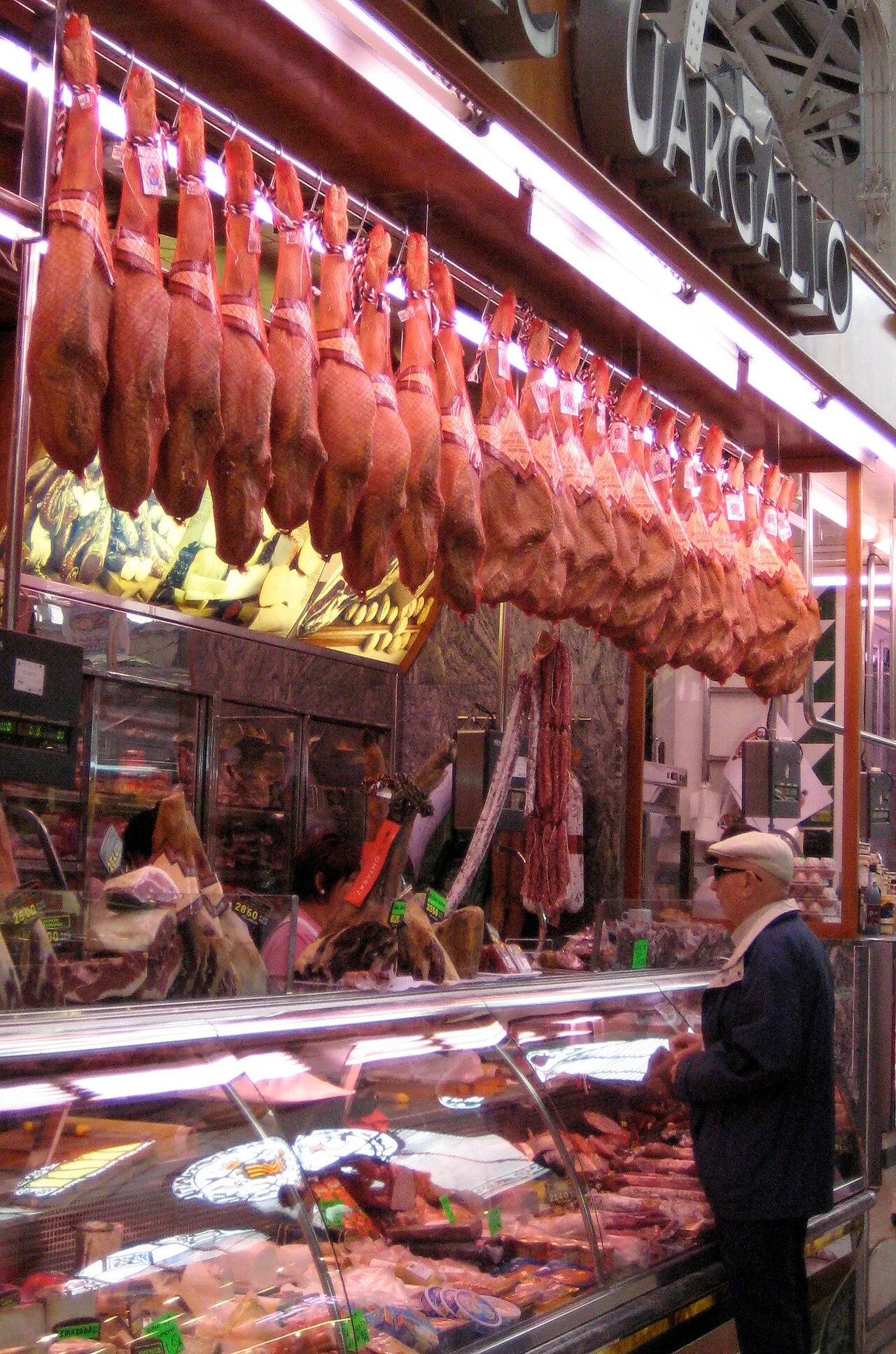 Vendedor atendiendo a un cliente en un puesto de jamones del Mercado Central de Valencia, con piezas de jamón curado colgadas detrás del mostrador.