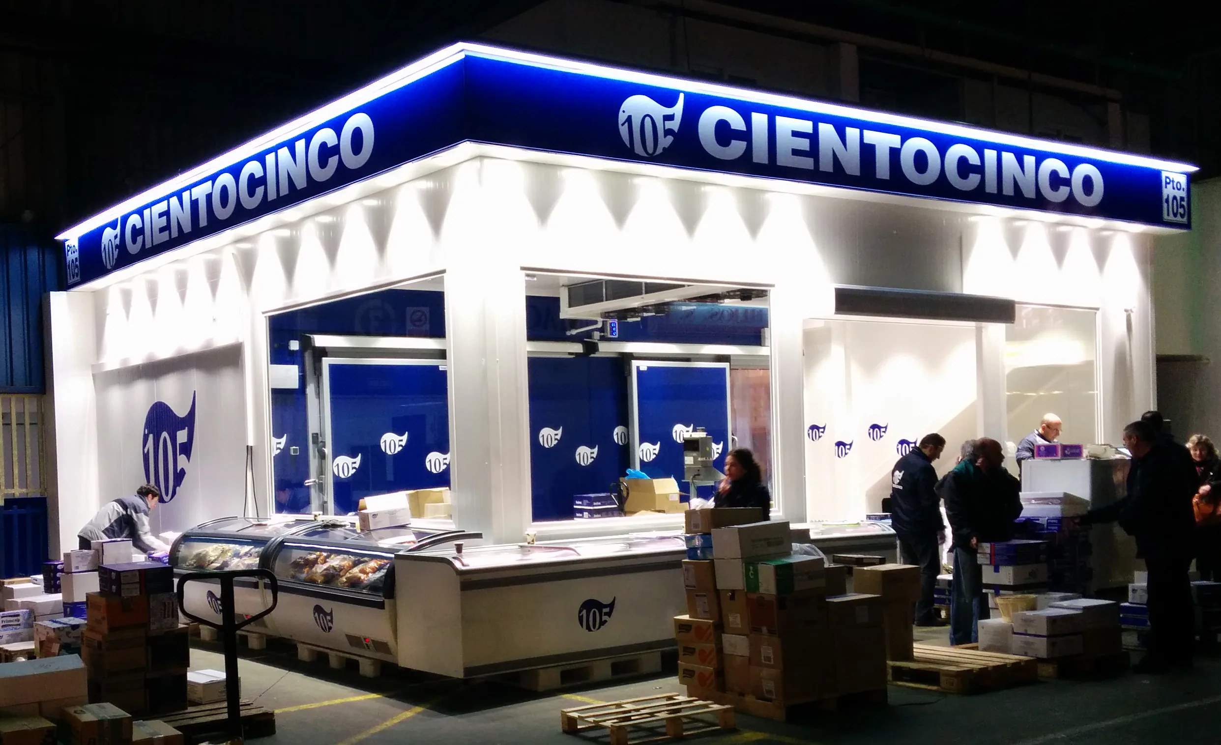 Puesto de venta de fruta y verdura en una nave mayorista de Mercamadrid, con cajas apiladas y precios visibles.