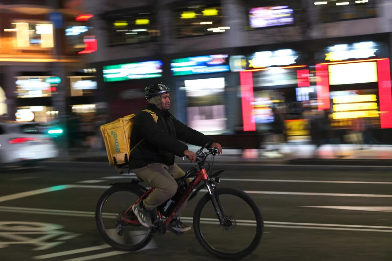 Repartidor de Glovo pedaleando de noche por una gran avenida, con la mochila amarilla a la espalda y los escaparates desenfocados por el movimiento.