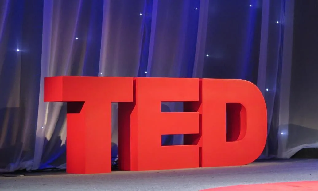 Ponente en un escenario TED con el icónico círculo rojo del logo iluminado al fondo.
