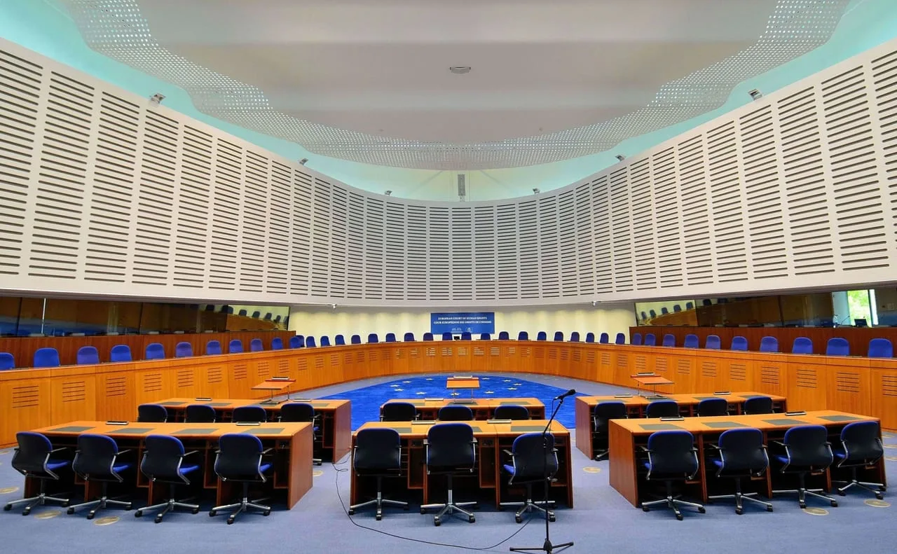 Sala de vistas vacía del Tribunal Europeo de Derechos Humanos en Estrasburgo, con el estrado en semicírculo y el rótulo del tribunal al fondo.
