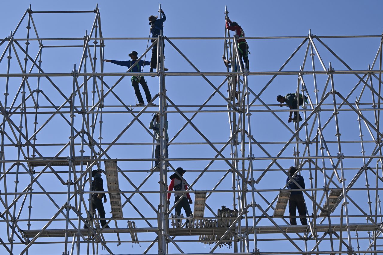 Trabajadores con casco, arnés y ropa de alta visibilidad montando un andamio metálico.