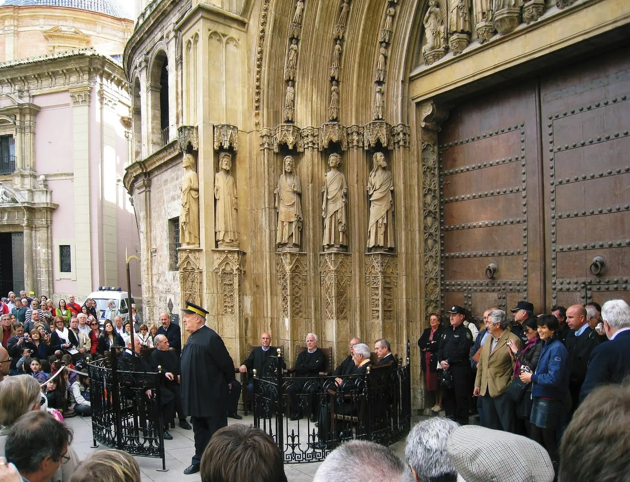 Sesión del Tribunal de les Aigües ante la puerta de los Apóstoles de la catedral de València, con los síndicos sentados dentro del recinto de hierro y el público alrededor.