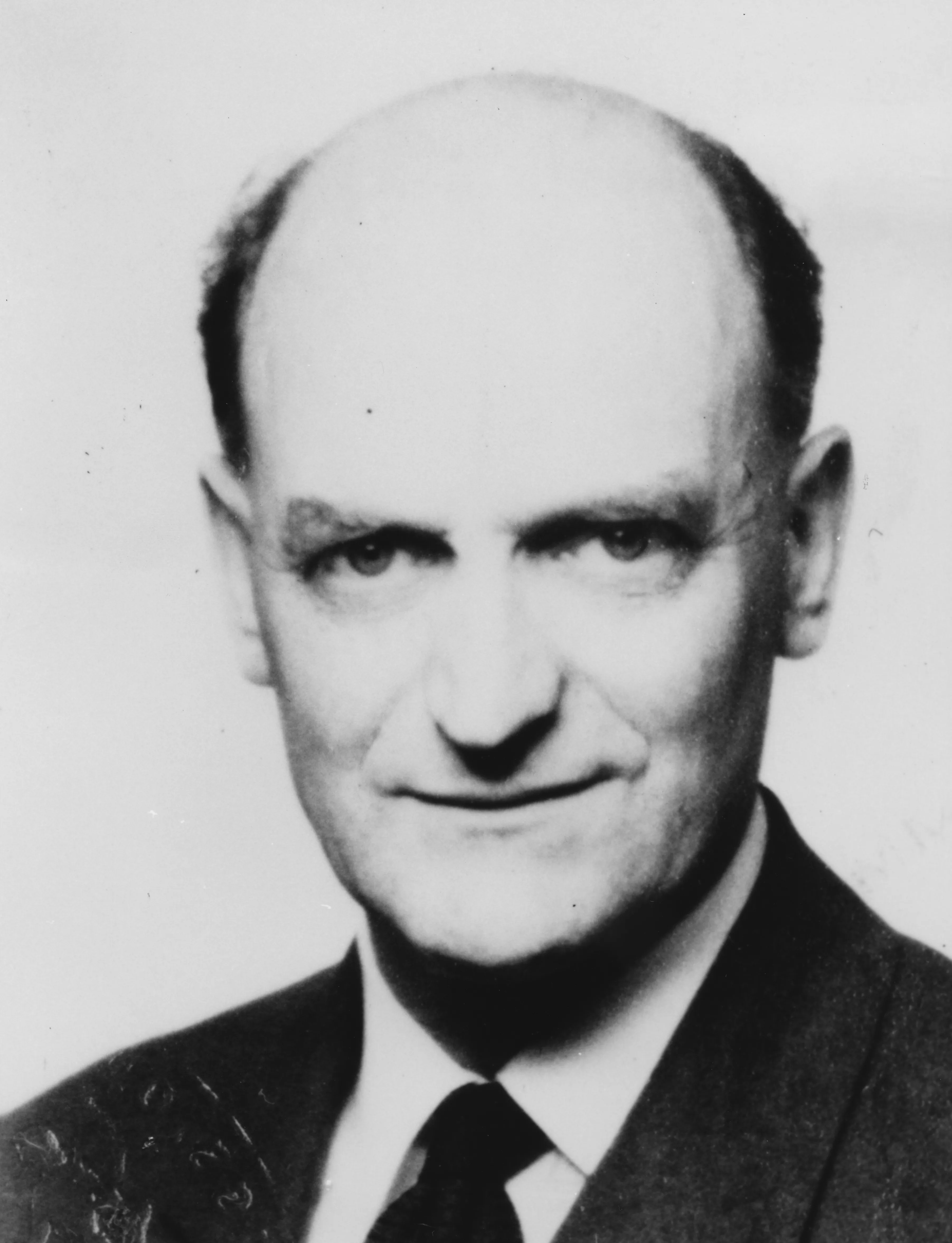 Retrato fotográfico en blanco y negro de A. W. H. Phillips (1914-1975), economista neozelandés profesor de la London School of Economics, sentado en una butaca con traje y corbata.